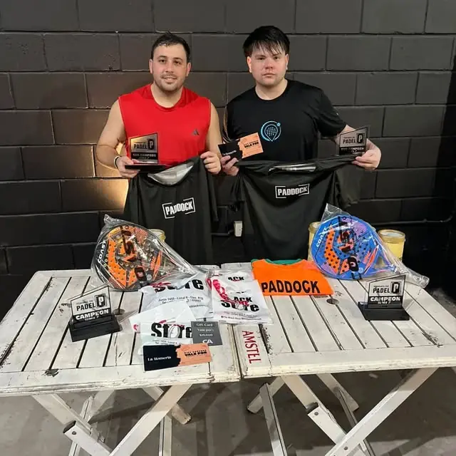 Pareja ganadora de un torneo jugado en Paddock, frente a la pared negra, con trofeos, paletas y remeras Paddock